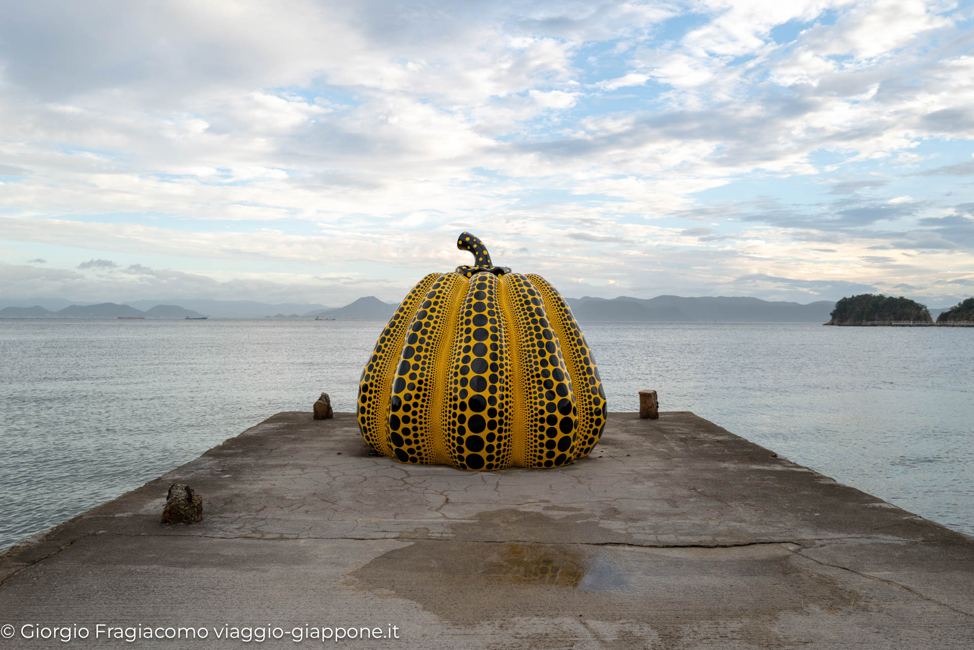 "Scultura di zucca gialla e nera sul molo, con vista sul mare e sulle montagne giapponesi sullo sfondo."