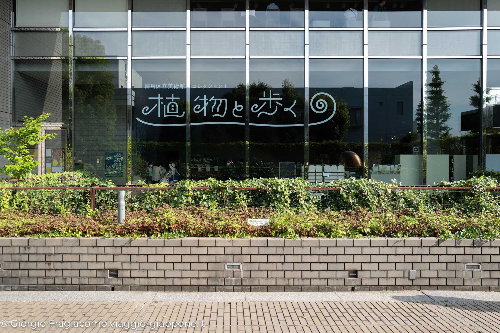 Vetrina di un museo giapponese con scritte artistiche e giardini curati, incorniciata da piante verdi.