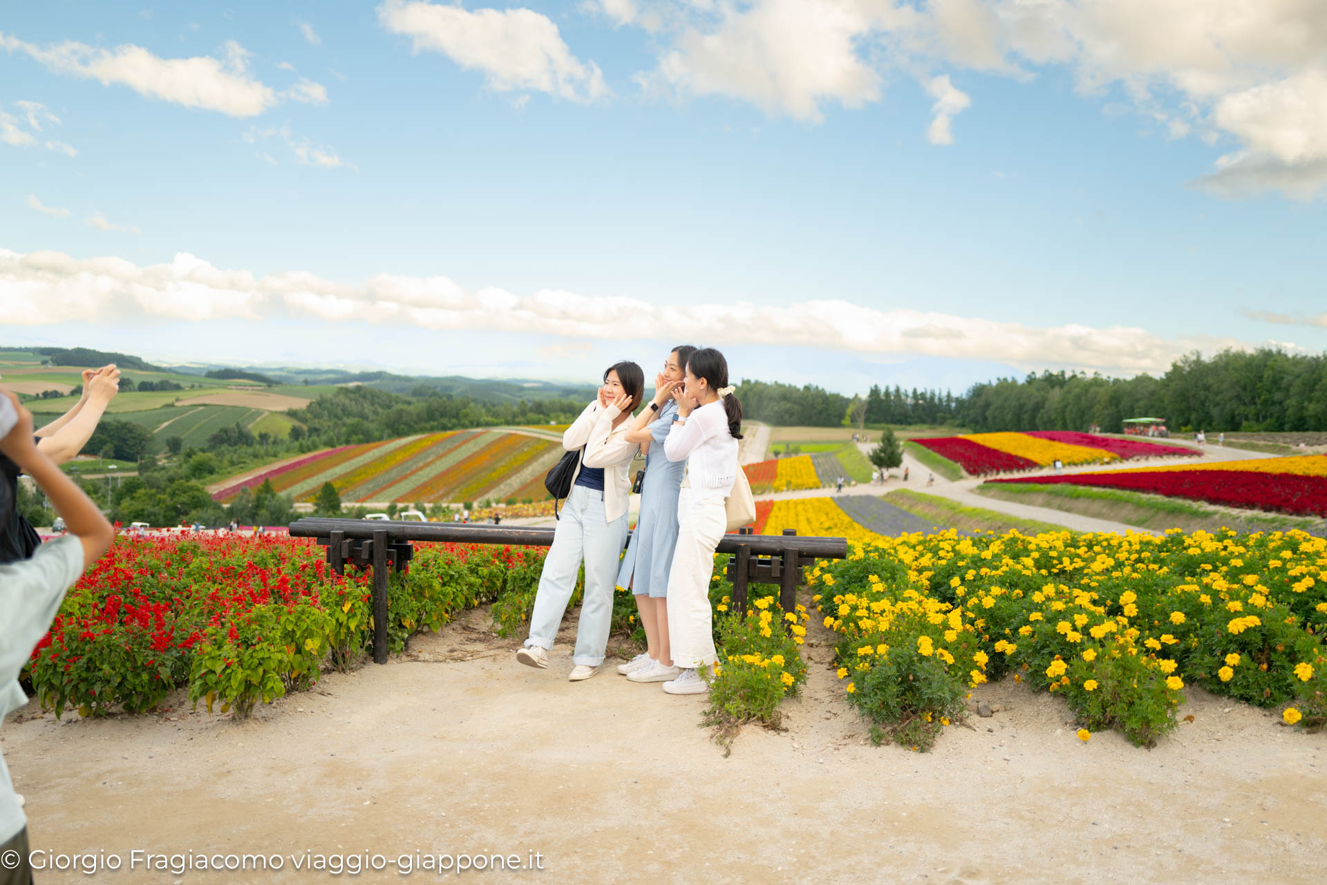 Tre ragazze sorridenti posano tra i fiori colorati in un campo giapponese, con un panorama mozzafiato sullo sfondo.