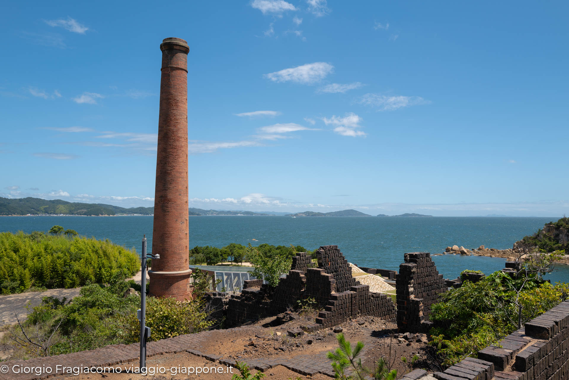 Ruderi di un'antica fabbrica con camino, vista panoramica sul mare e colline giapponesi sullo sfondo.