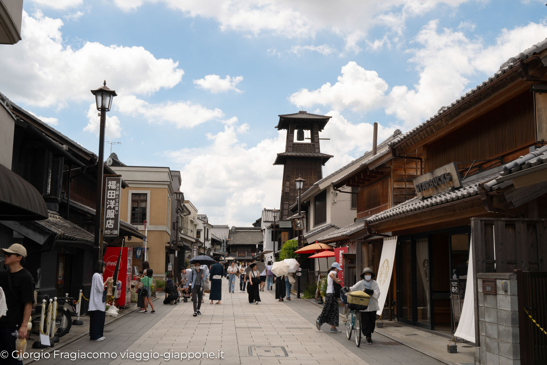 Strada tradizionale giapponese con negozi, gente in kimono e un tipico campanile in legno sullo sfondo.