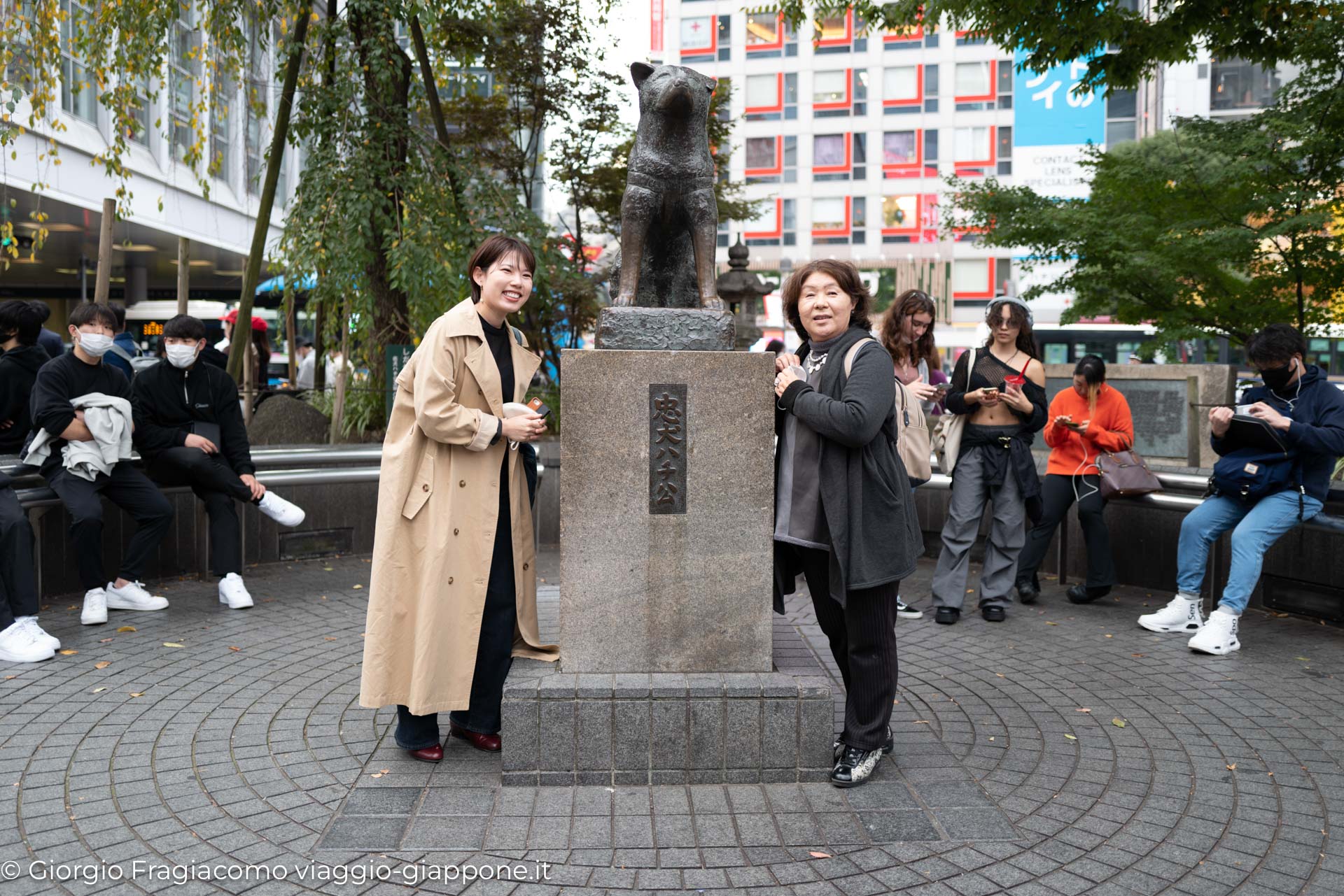 Due donne sorridenti posano accanto alla statua di Hachiko, simbolo di fedeltà, in Giappone.